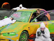 schauspielende in Maleroveralls stehen rund um ein bunt bemaltes Auto