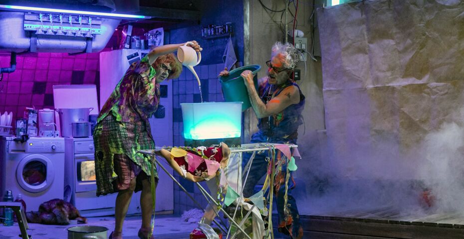 Zwei Schauspielende in bunten zerrissenen Kostümen gießen Flüssigkeiten in ein leuchtendes Gefäß