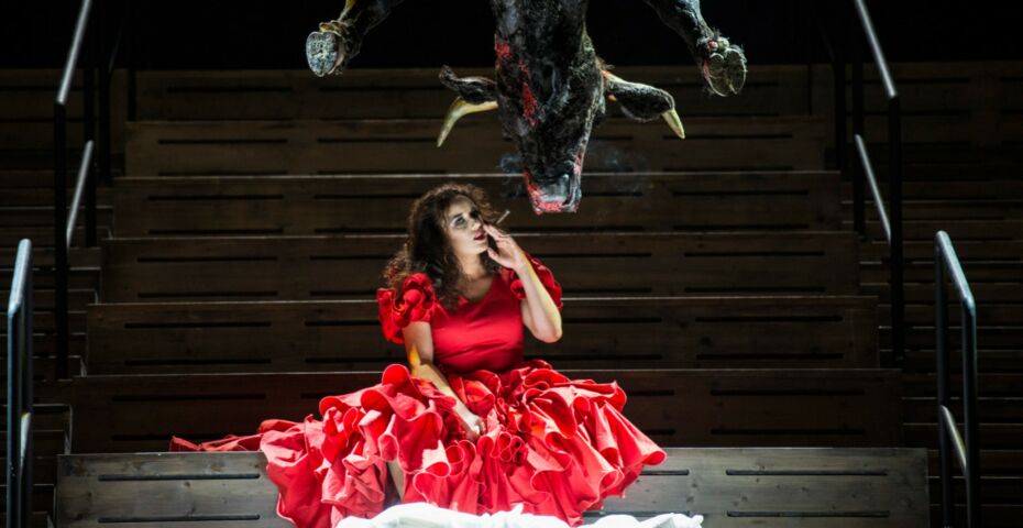 Eine Frau mit langen dunklen gelockten Haaren sitzt in rotem Kleid auf einer Bühne und hält ihre linke Hand ans Gesicht, von oben hängen Tierkadaver herunter