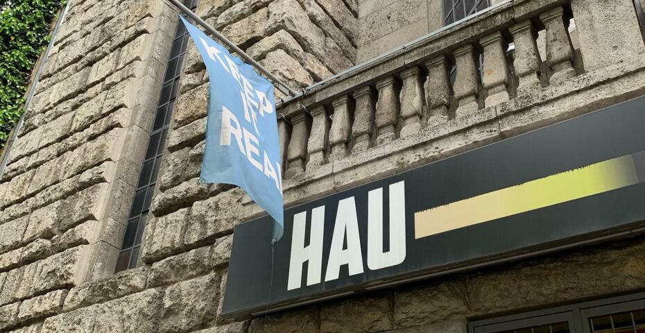 Die Fassade des Hau, der Eingang ist von Innen beleuchtet, darüber eine blaue Fahne mit der Aufschrift "Keep it real"