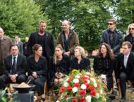 Hinter einem Begräbnis mit Blumenstrauß sitzen und stehen in zwei Reihen schwarz gekleidete Männer und Frauen.