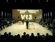 Auf einer Leinwand der Schriftzug V13 vor einem barocken Gebäude.