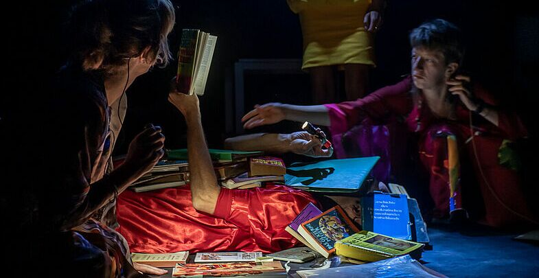 Eine Person im roten Schlafanzug liegt auf dem Rücken auf  der Bühne und ist mit Büchern und Comics bedeckt. Links und Rechts neben ihr andere Darstellerinnen mit ausgebreiteten Händen.