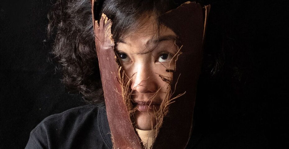 Vor schwarzem Hintergrund eine Person mit gelockten Haaren hält sich zwei Holzstücke vor das Gesicht, zwischen denen sie hindurch schaut.