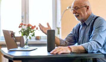 Ein älterer Mann sitzt an einem Tisch und bedient per Sprachsteuerung einen Smartspeaker