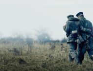 Zwei Soldaten in historischen Uniformen stützen sich gegenseitig auf dem Weg über eine Wiese unter grauem Himmel