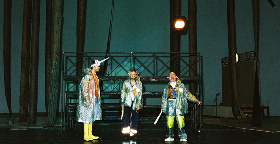 Drei Schauspieler in silbernen Regencapes stehen auf einer Bühne und schneiden Grimassen