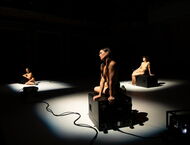drei unbekleidete Schauspielerinnen sitzen in Scheinwerferlicht auf einer ansonsten Dunklen Bühne auf schwarzen Kisten