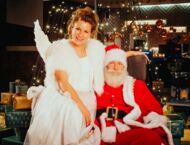 Ein Mann im Weihnachtsmannkostüm sitzt in einem Sessel vor einem geschmückten Baum. Auf seiner Armlehne sitzt eine Frau in einem Engelsskostüm. Beide lächeln in die Kamera.
