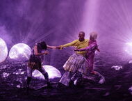 bunt gekleidete Tänzerinnen stehen in violettem Licht auf einer Bühne