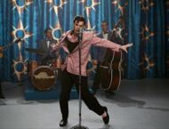 Elvis in Tanzpose, seine Arme sind ausgestreckt, sein linkes Bein zeigt nach links, vor ihm ein Mikrofon und hinter ihm seine Band