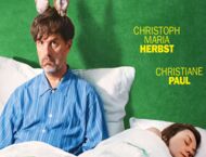 Vor einer grünen Wand sitzt ein Mann aufrecht im Bett, neben ihm schläft eine Frau ihm abgewandt