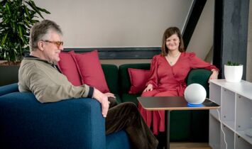 Unter einem Schrägdach sitzt links ein älterer lächelnder Mann in einem blauen Sessel und rechts eine junge Frau mit Seitenscheitel und offenem Haar, dazwischen auf einem Tisch eine weiße Kugel