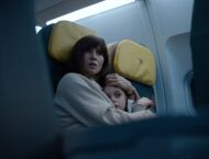 Eine junge Frau mit dunklen Haaren und Pony sitzt in einem Flugzeug und hält ihren Sohn eng im Arm, ihr Mund ist leicht geöffnet und ihr Blick angsterfüllt.