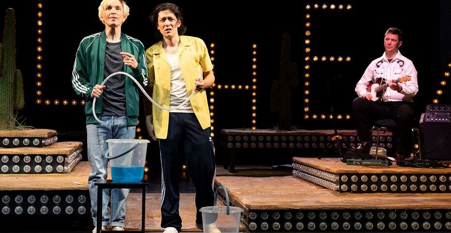 2 SChauspielende stehen ratlos vor einem Eimer mit blauer Flüssigkeit. Im Hintergrund ein Mann mit einer E-Gitarre auf einem Barhocker