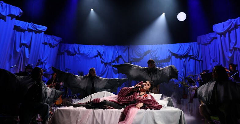 Eine Schauspielende liegt in Robe auf einem Bett. Um sie herum Vögel