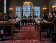 Im repräsentativen Besprechungssaal sitzen die Teilnehmer an einem Tisch in U-Form. Auf dem Tisch liegen Besprechungsunterlagen. Auf Tabletts stehen kristallene Gläser und Wasserkaraffen. Die Teilnehmer wenden ihre Blicke zu Reinhard Heydrich.