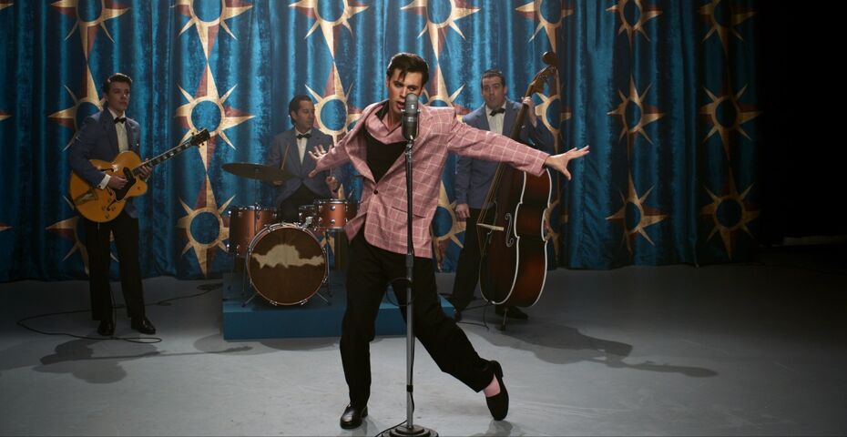 Elvis in Tanzpose, seine Arme sind ausgestreckt, sein linkes Bein zeigt nach links, vor ihm ein Mikrofon und hinter ihm seine Band