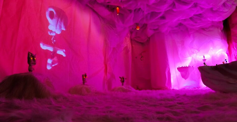 Ein in violettes Licht getauchter Raum beinhaltet verschiedene Puppen mit leuchtenden Augen